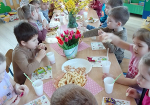 Dzieci siedzą przy stole, na którym są: wazony z wiosennymi kwiatami, talerze z chrupkami i słodyczami, kubeczki ze słomkami.