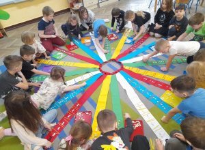 Na dywanie rozłożony jest Wiatrak matematyczny. Dzieci układają wzór z patyczków i fasolek na wstążce, przed którą siedzą.