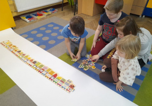 Czworo dzieci siedzi na dywanie i układa matematyczny pociąg na białym kartonie.