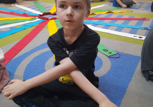 Dzieci siedzą na dywanie odwrócone tyłem do Wiatraka matematycznego. Oskar mówi jaki kolor ma jego wstążka, jaka cyfra na niej się znajduje i jaki znaczek.