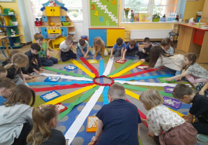 Dzieci siedzą wokół Wiatraka matematycznego na dywanie. Przedszkolaki układają na swoich wstążkach fasolki zgodnie z umieszczonymi na nich cyframi.