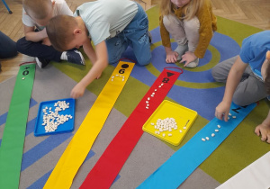Czworo dzieci podczas zabawy z Wiatrakiem matematycznym układa na wstążce przy której siedzi o 1 więcej fasolkę.