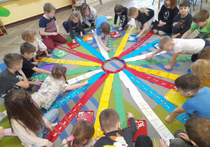 Wiatrak rozłożony jest na dywanie. Dzieci układają kolejny wzór z patyczków i fasolek na wstążce, przed którą siedzą.