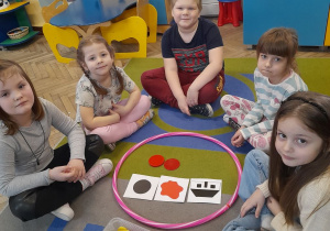 Zabawa klockami Dienesa - pięcioro dzieci siedzi na dywanie przy obręczy. Przedszkolaki wybrały klocki zgodnie z kodem obrazkowym - duże, czerwone koła.