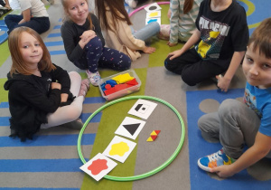 Winicjusz, Gabrysia, Oskar i Karol siedzą przy obręczy, do której włożyli małe, czerwone i żółte klocki w kształcie trójkątów. Za nimi inna grupka dzieci wybiera klocki według kodu.