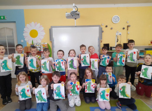 Dzieci prezentują namalowane portrety Dinka - maskotki programu "Czyste powietrze wokół nas". W tle tablica multimedialna, kącik książki.
