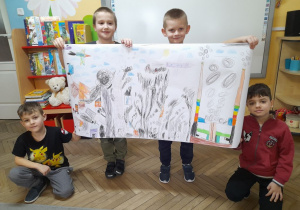 Eryk, Franek, Kacper i Tymon trzymają wykonany przez siebie plakat przedstawiający różne rodzaje dymów. W tle tablica multimedialna, kącik książki.