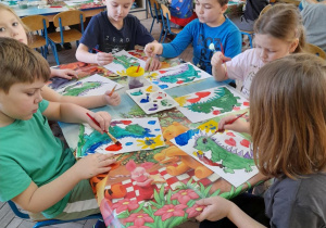 Dzieci siedzą przy stołach i malują farbami plakatowymi portret Dinka - maskotki programu "Czyste powietrze wokół nas". W tle szafki z układankami.