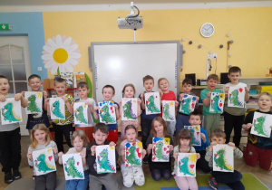 Dzieci prezentują namalowane portrety Dinka - maskotki programu "Czyste powietrze wokół nas". W tle tablica multimedialna, kącik książki.