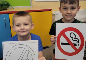 Kacper i Eryk siedzą na dywanie. Chłopcy trzymają w dłoniach znaki "Zakaz palenia".