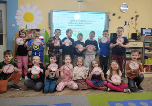 Przedszkolaki stoją przed tablicą multimedialną. Dzieci trzymają w dłoniach pomalowane przez siebie znaki "Zakaz palenia".