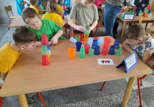 Chłopcy siedzą przy stole i układają wieże z kolorowych kubków według podanego wzoru.