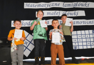 Karol, Arek, Eryk i Filip pozują do zdjęcia z otrzymanymi dyplomami i nagrodami. W tle napis - Matematyczne łamigłówki dla małej główki - Od kodowania do programowania.