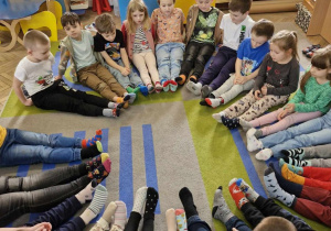 Przedszkolaki siedzą w kółeczku na dywanie. Wszystkie dzieci mają zdjęte buciki by pokazać kolorowe skarpetki, które założyły tego dnia na znak solidarności z osobami z zespołem downa.
