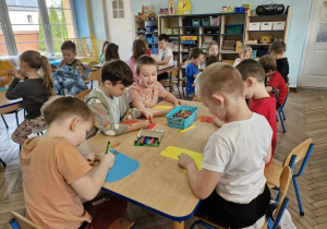 Filip, Tymek, Franek, Antoś i Kacper kolorują papierowe skarpetki siedząc przy stoliczku. W tle widać pozostałe dzieci z grupy.