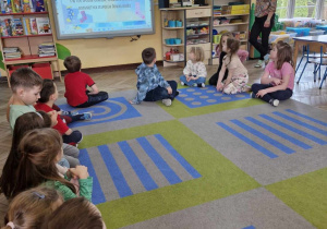 Dzieci siedzą na dywanie i oglądają prezentację multimedialną, która przybliża im zagadnienia związane z zespołem downa. W tle widać panią pedagog specjalną Edytę.