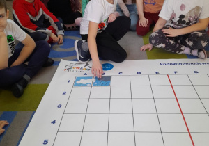Dzieci siedzą obok maty na dywanie, a Kacper układa element obrazka na podstawie podanych koordynatów.