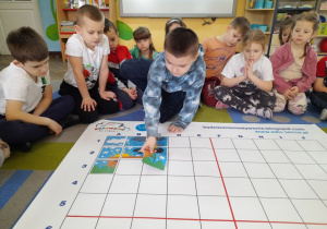 Dzieci siedzą na dywanie, a Gabryś dokłada kolejny element do obrazka na podstawie podanych koordynatów.