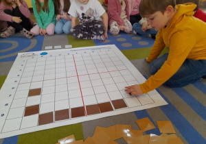 Kacper na podstawie podanych koordynatów układa na macie brązowe kwadraty, a koleżanki i koledzy obserwują działanie chłopca.