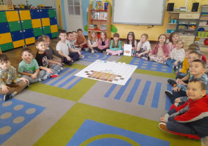 Dzieci siedzą na dywanie wokół maty do kodowania, na której znajduje się ułożony obrazek garnka z brązowych kwadratów i symbole zjawisk atmosferycznych.