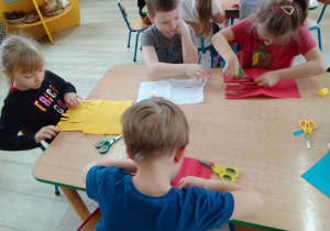 Dzieci przy stolikach wycinają nożyczkami po linii paski kolorowego papieru.