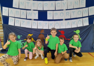 Sześcioro dzieci z drużyny "Stokrotki", ubranych w zielone koszulki, przykucnęło przed dekoracją, na której umieszczony jest napis "Integracyjne Zabawy Sportowe Przedszkolaków".