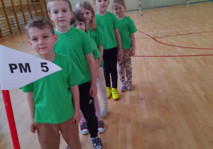 Dzieci ubrane w zielone koszulki stoją w rzędzie jedno za drugim przed swoim stanowiskiem na sali gimnastycznej.