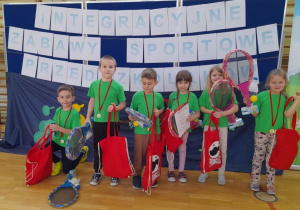 Dzieci pozują do zdjęcia z otrzymanymi nagrodami. W tle napis "Integracyjne Zabawy Sportowe Przedszkolaków"