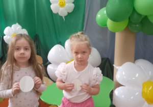Tosia i Amanda prezentują swoje ekologiczne kule. W tle zielony materiał, na którym przypięte są białe balonowe stokrotki oraz drzewo wykonane z zielonych balonów.