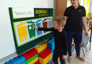 Antek na tablicy przypina butelkę do sylwety pojemnika w zielonym kolorze. Obok przedstawicielka firmy PreZero Service Centrum.