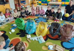 Na dywanie worki na śmieci w różnych kolorach i rozrzucone śmieci, wokół siedzą dzieci.