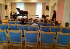 Na scenie dwie uczennice Szkoły Muzycznej grają na fortepianie, a przedszkolaki siedzą na krzesłach. Obok dzieci stoi nauczyciel szkoły.