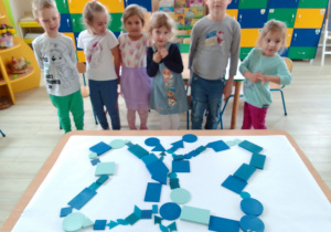 Grupa dzieci prezentuje ułożonego z niebieskich figur geometrycznych motyla