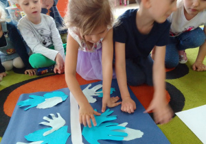 Matylda przykleja papierową dłoń do sylwety motyla. Wokół inne dzieci obserwują jej działanie.