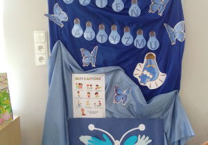 Dekoracja z napisem Dzień Autyzmu, ozdobiona pracą plastyczną wykonaną przez dzieci – niebieskim motylem z papierowych dłoni.