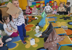 Dzieci siedzą na dywanie, a w środku stoi Samuel z kartonowym pudełkiem. Obok znajdują się worki w różnych kolorach i rozrzucone śmieci.