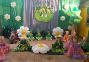 Dzieci przebrane za motylki, stokrotki, wiewiórki, ptaszki, jeżyki, zielone skrzaty siedzą na tle dekoracji.