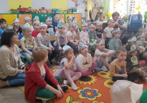 Przedszkolaki siedzą na dywanie ze swoimi paniami. Dzieci mają na głowach opaski z okazji Dnia Ziemi.