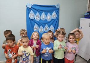 Dzieci z grupy "Pszczółki" stoją na tle dekoracji trzymając przed sobą otrzymane odznaki z napisem "Przyjaciel wody".