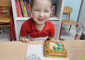 Uśmiechnięty Szymon pozuje do zdjęcia, przed nim na serwetce leży kolorowa ciasteczkowa pisanka i udekorowany mazurek.