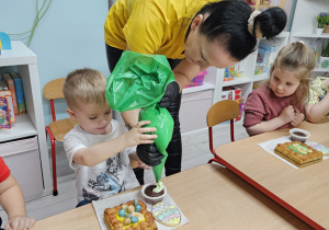 Ignaś wspólnie z panią dekoruje swoją muffinkę jasnozielonym kremem.