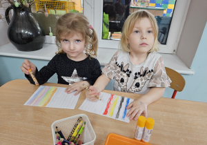 Maja S. i Nadia siedzą przy stoliku i kolorują kartki papieru.