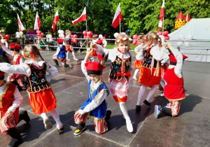 Dzieci ubrane w stroje krakowskie tańczą na scenie w parku Traugutta. Chłopcy klęczą na kolanie z ręką podniesioną do góry, a dziewczynki podskakują wokół nich.