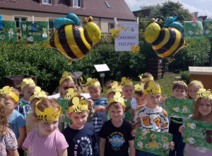 Dzieci z grupy "Motylki" prezentują wykonane przez siebie prace - łąka dla pszczółek. Nad dziećmi wiszą dwie pszczoły oraz napis "Chrońmy pszczoły".