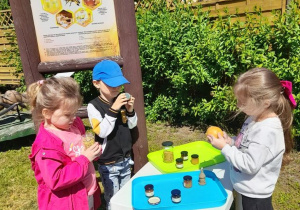 Czworo dzieci z grupy "Motylki" stoi obok stolika, na którym znajdują się produkty pszczele. Obok tablica edukacyjna "Co nam daje pszczoła".