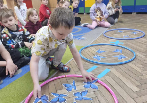 Przedszkolaki siedzą na dywanie. Przed nimi leżą trzy obręcze, w których znajdują się niebieskie motyle. Dzieci wykonują działania matematyczne.