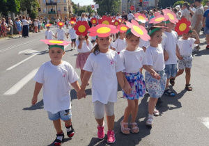 Dzieci w białych koszulkach i kwiatowych nakryciach głowy idą ulicą podczas Parady Kwiatowej. W tle widownia, budynki, drzewa.