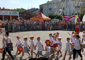 Dzieci z grupy "Słoneczka" razem z nauczycielami maszerują w Paradzie Kwiatowej. W tle widownia, budynki, karuzela - diabelski młyn.