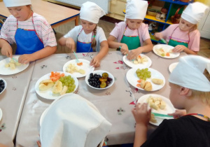 Grupa dziewczynek w kucharskich strojach drobni plastikowymi nożami owoce. Przed nimi na stole stoją talerze z owocami.