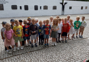 Dzieci stoją przed tablicami pamiątkowymi umieszczonymi na "Murze pamięci". Na murze znajduje się napis - Muzeum Bitwy nad Bzurą w Kutnie. Chłopiec z dziewczynką trzymają biało-czerwone znicze.